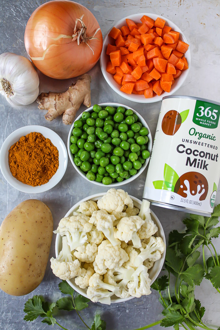 Onion, garlic, ginger, carrots, peas, cauliflower, potato, and coconut milk ingredients laid out on a metal tray