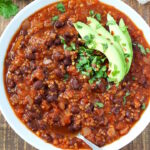 Smoky chipotle black bean quinoa chili in a white bowl with avocado and serving spoon