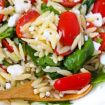 Close up view of orzo spinach feta salad with tomato on a white plate with bamboo serving spoon