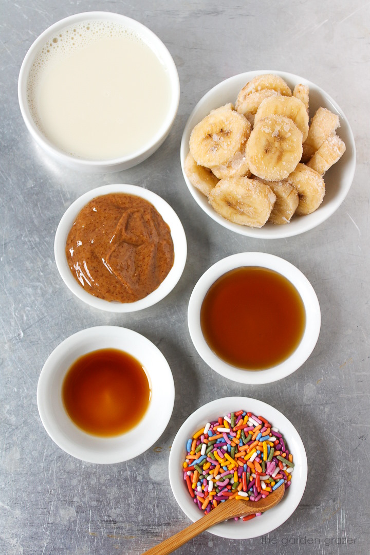 Soy milk, almond butter, frozen banana, pure maple syrup, vanilla, and sprinkle ingredients laid out on a metal tray