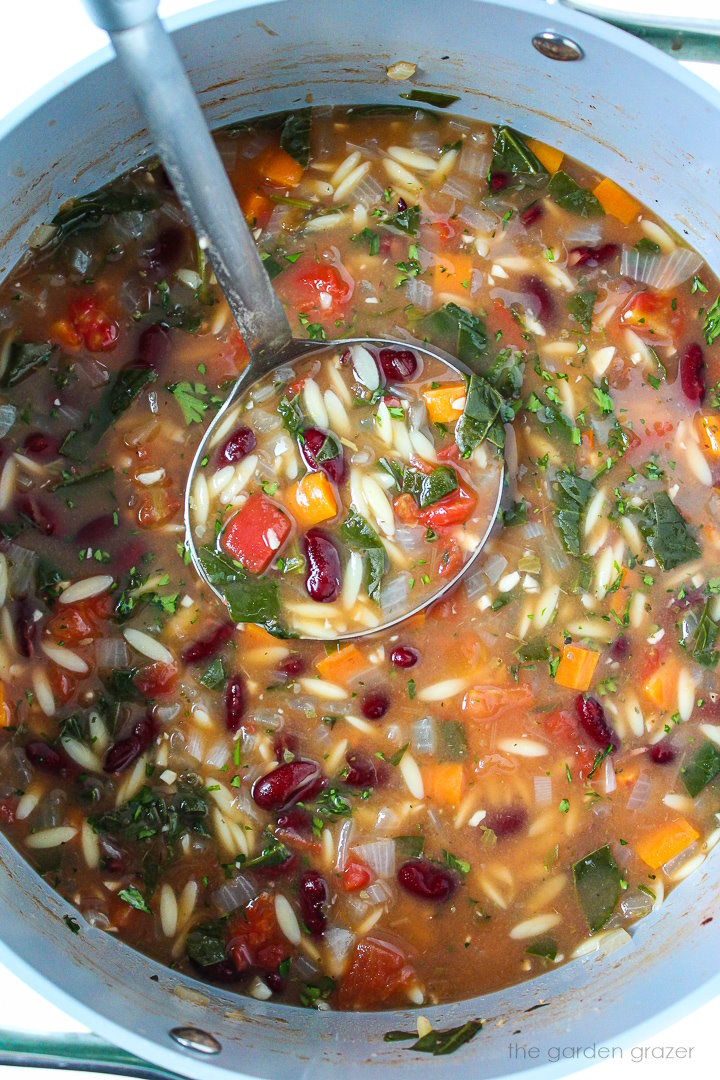 A ladle lifting up kidney bean orzo soup as it cooks in a large stockpot 