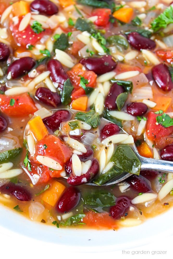 Close-up view of a spoon lifting up some kidney bean, spinach, and orzo soup from a white bowl