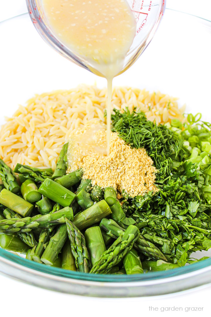 Pouring the dressing over asparagus orzo in a large glass bowl