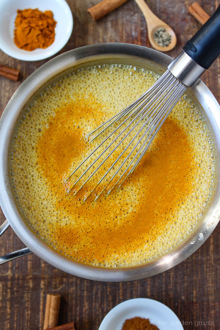 Overhead view of turmeric latte heating in a small pan with a metal whisk