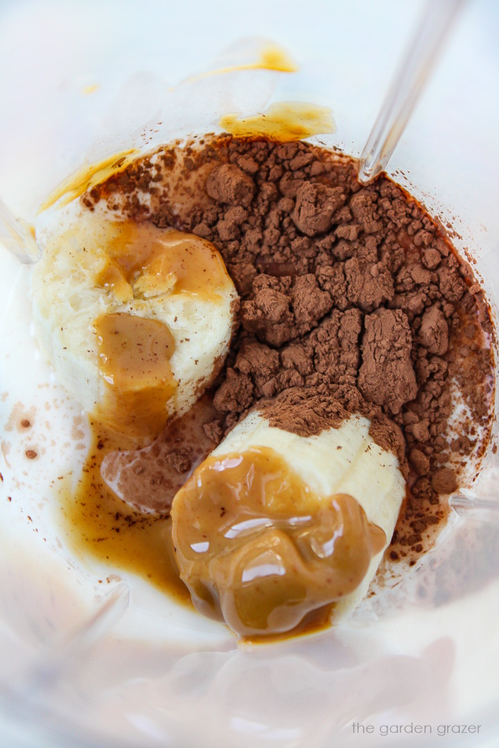 Overhead view of banana, cacao powder, milk, and peanut butter in a blender cup before blending