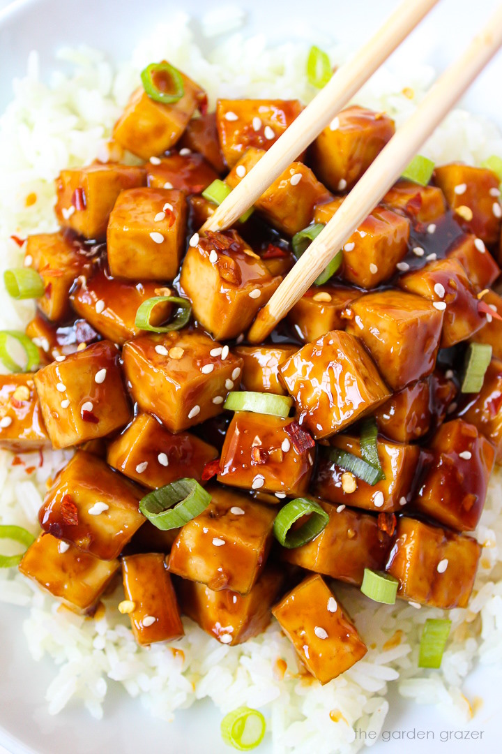 Chopsticks picking up teriyaki baked tofu from a white bowl