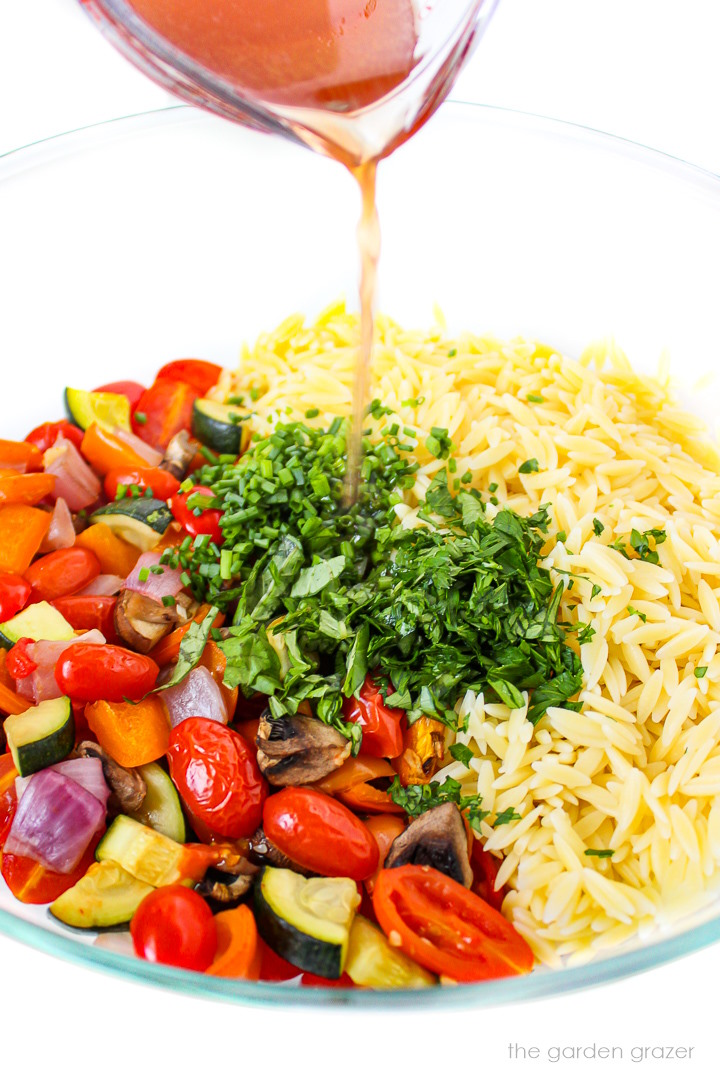 Lemon vinegar dressing being poured over a bowl of orzo, herbs, and roasted vegetables
