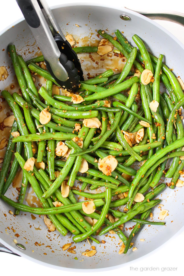 Tongs stirring green beans in a large skillet with garlic, almonds, and tamari.