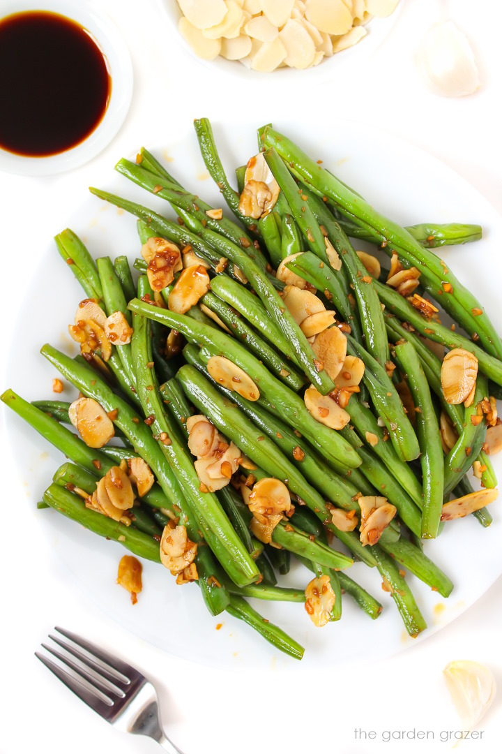 Cooked green beans with almonds, garlic, and tamari on a white plate with serving fork