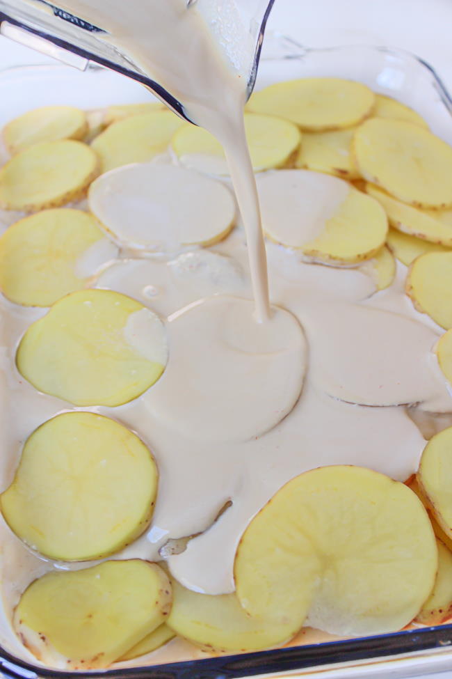 Cashew cheese sauce pouring on large dish of sliced potatoes