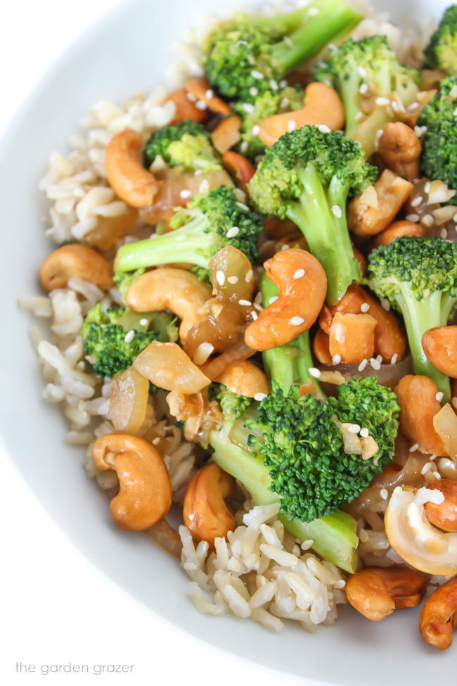 Nuts and veggies in a white bowl on top of cooked brown rice and sesame seeds