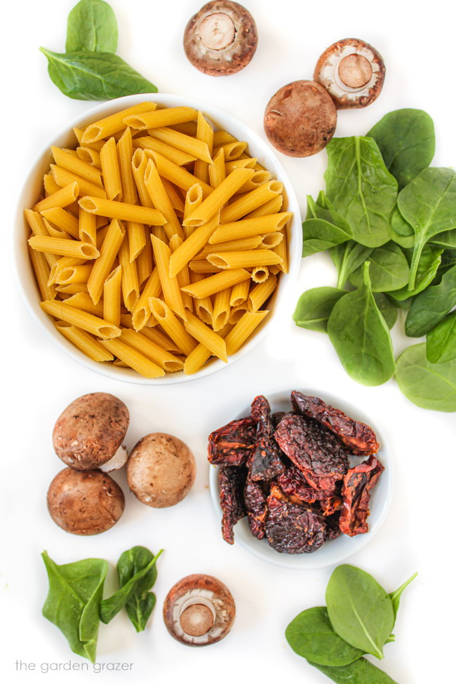 Dry penne pasta, spinach, tomatoes, and mushroom ingredients on a white table