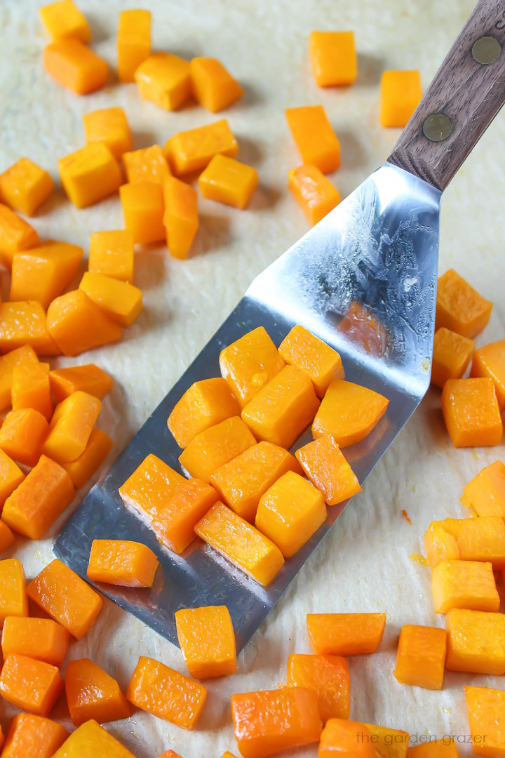 Metal spatula lifting up roasted butternut squash cubes from a sheet pan