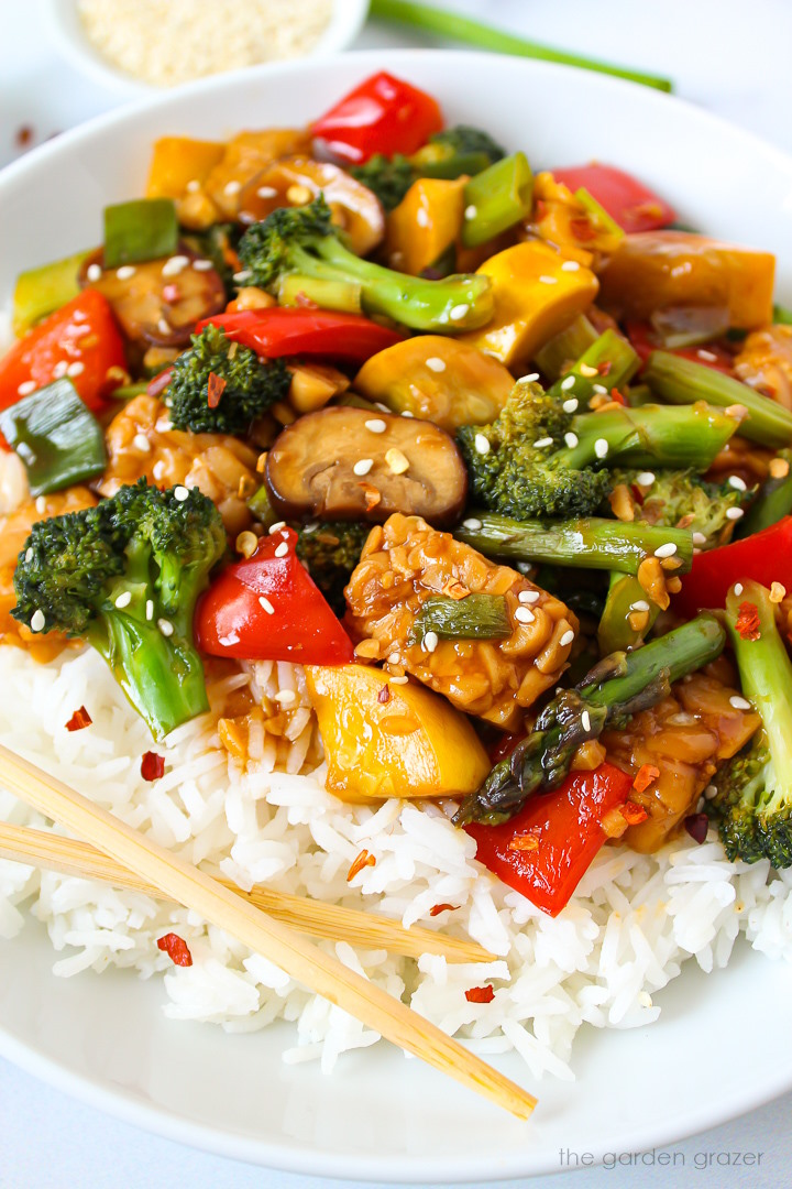 Close up view of teriyaki vegetable stir fry in a white serving bowl with rice and chopsticks