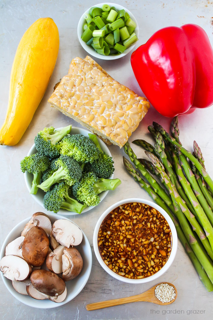 Red bell pepper, yellow squash, green onions, broccoli, asparagus, mushrooms, and sauce ingredients laid out on a metal tray