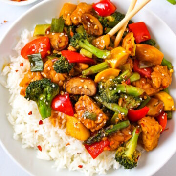 Teriyaki vegetable stir fry in a white serving bowl paired with rice and sesame seeds