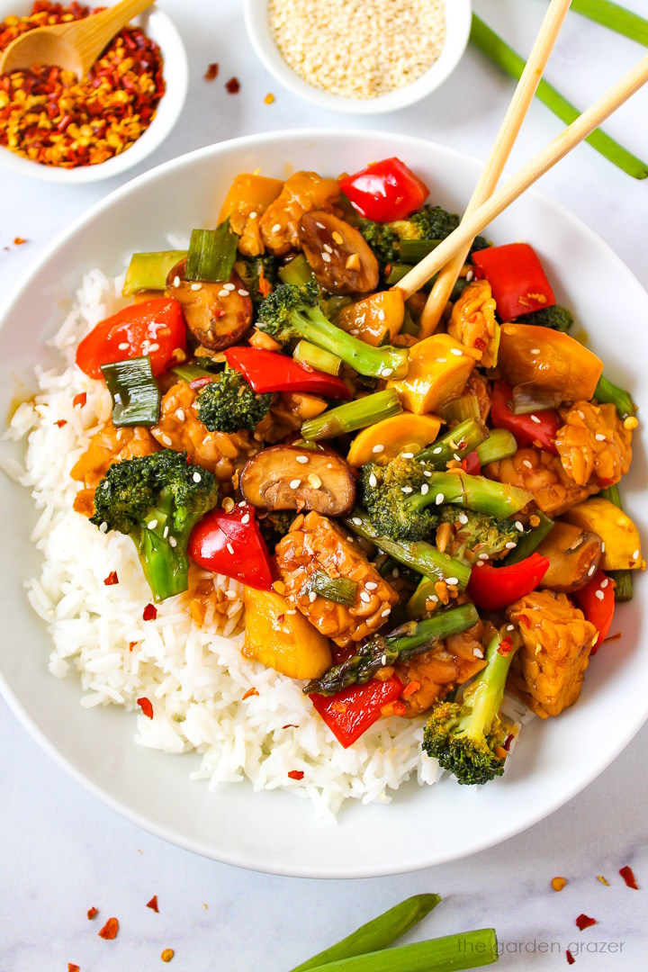 Teriyaki vegetable stir fry in a white serving bowl paired with rice and sesame seeds