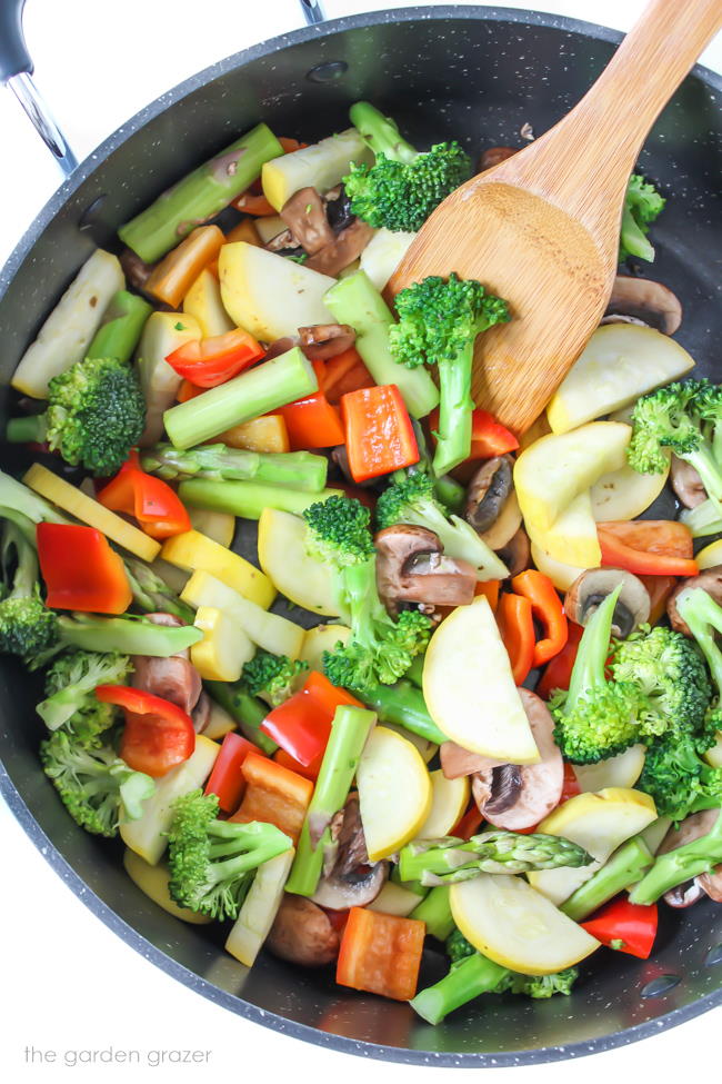 Vegetable stir fry cooking in a large skillet with wooden spatula