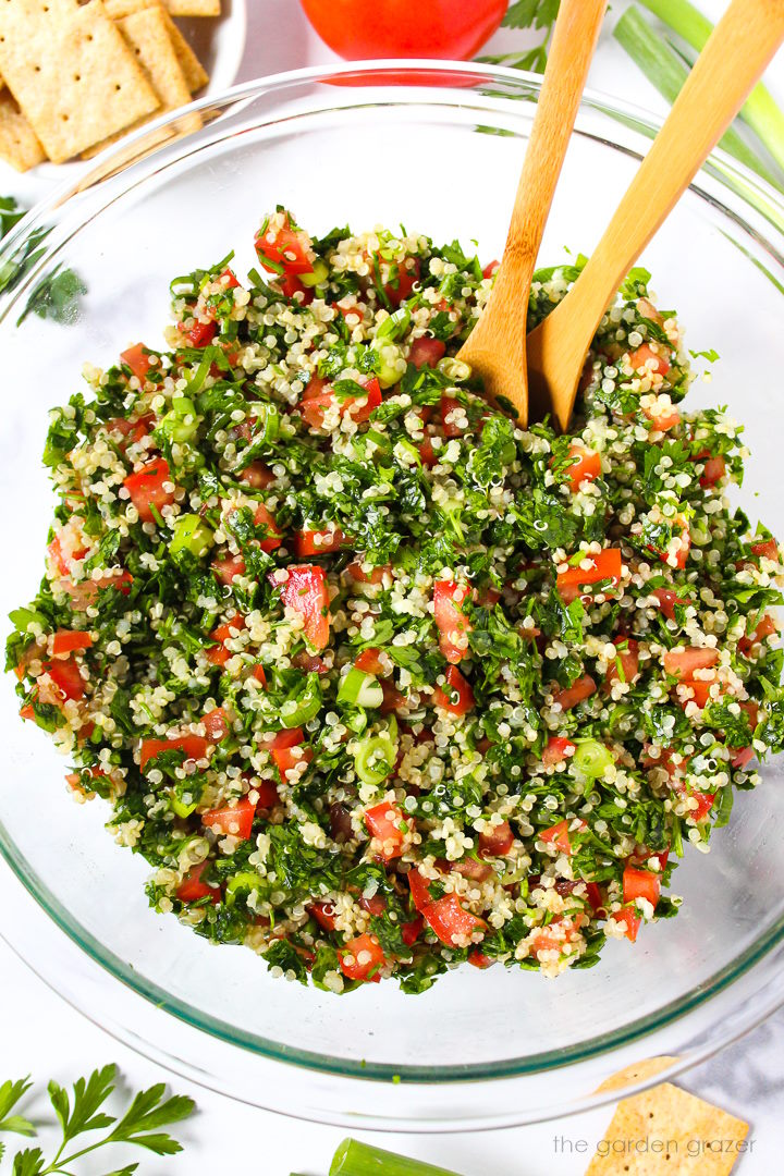 Overhead view of quinoa tabbouleh mixed together in a large glass bowl with wooden spoons
