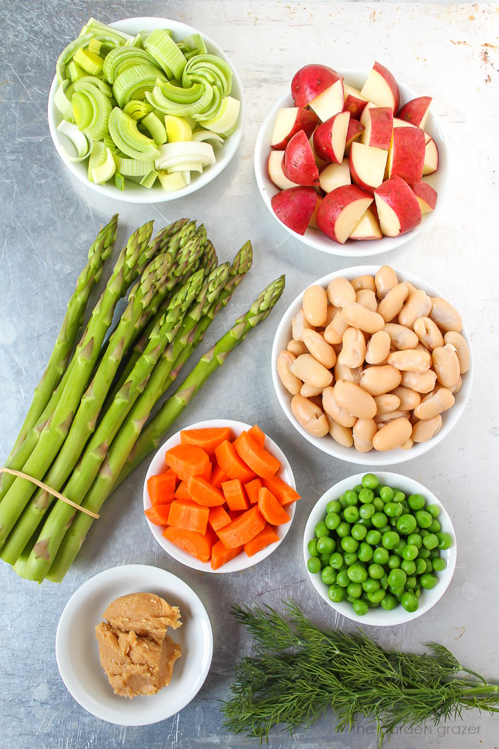 Fresh asparagus, white beans, leek, red potato, green peas, miso, and fresh dill ingredients laid out on a metal tray