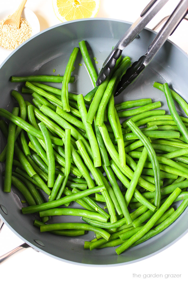 Overhead view of fresh beans cooking in a large skillet with tongs for stirring