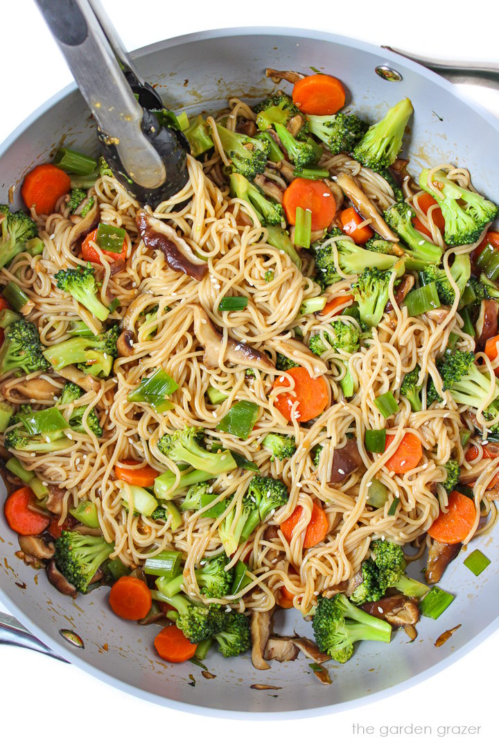Teriyaki noodles and vegetables cooking in a large skillet with tongs for stirring