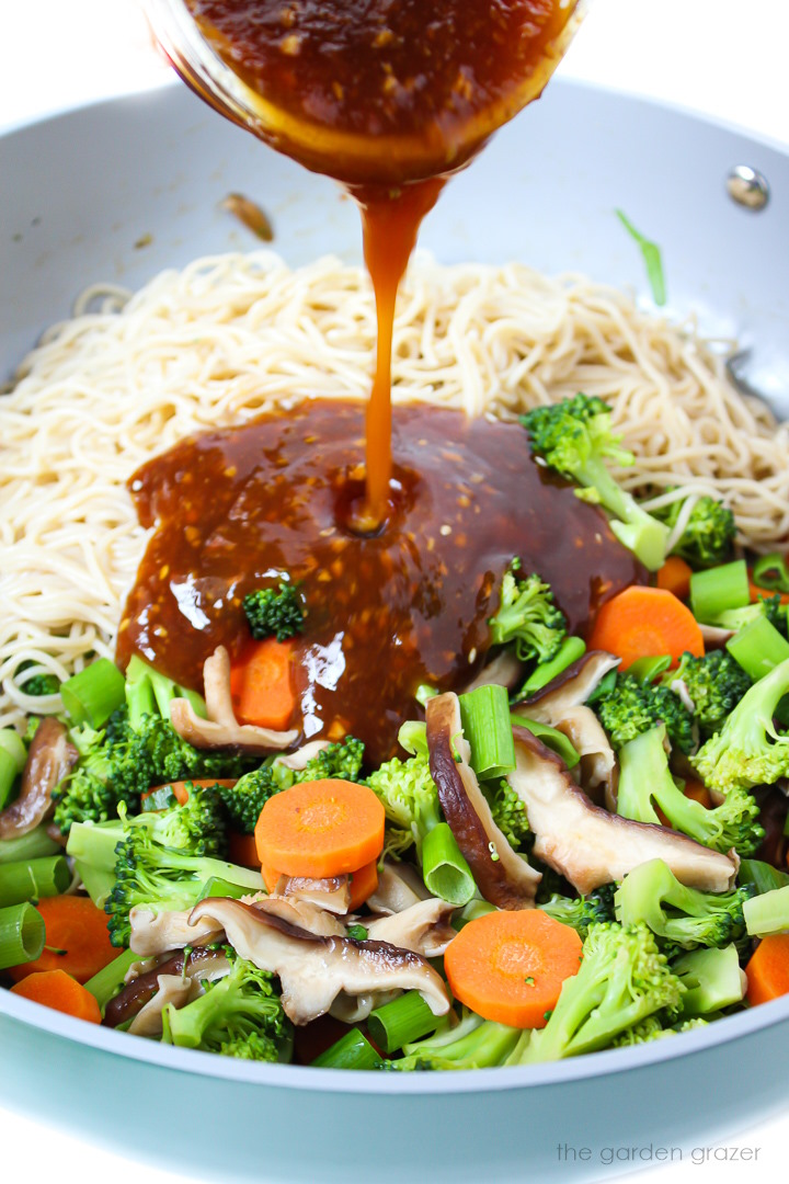 Teriyaki sauce being poured over noodles and vegetables in a large skillet