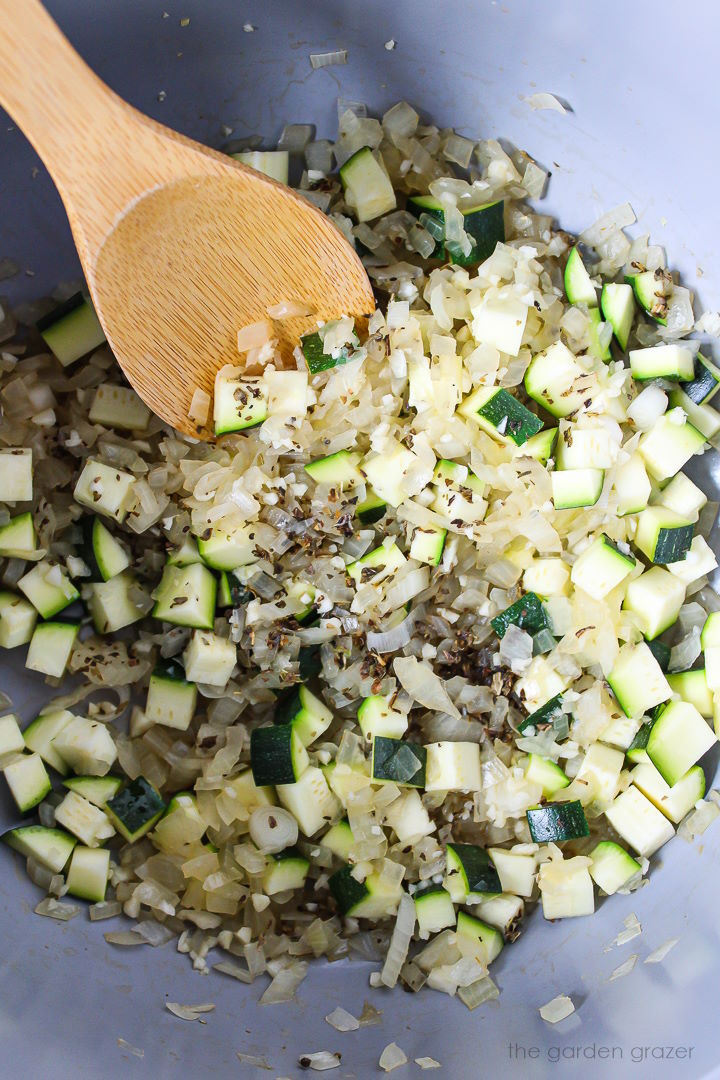 Onion, garlic, zucchini, and dried basil cooking in a stockpot with stirring spoon