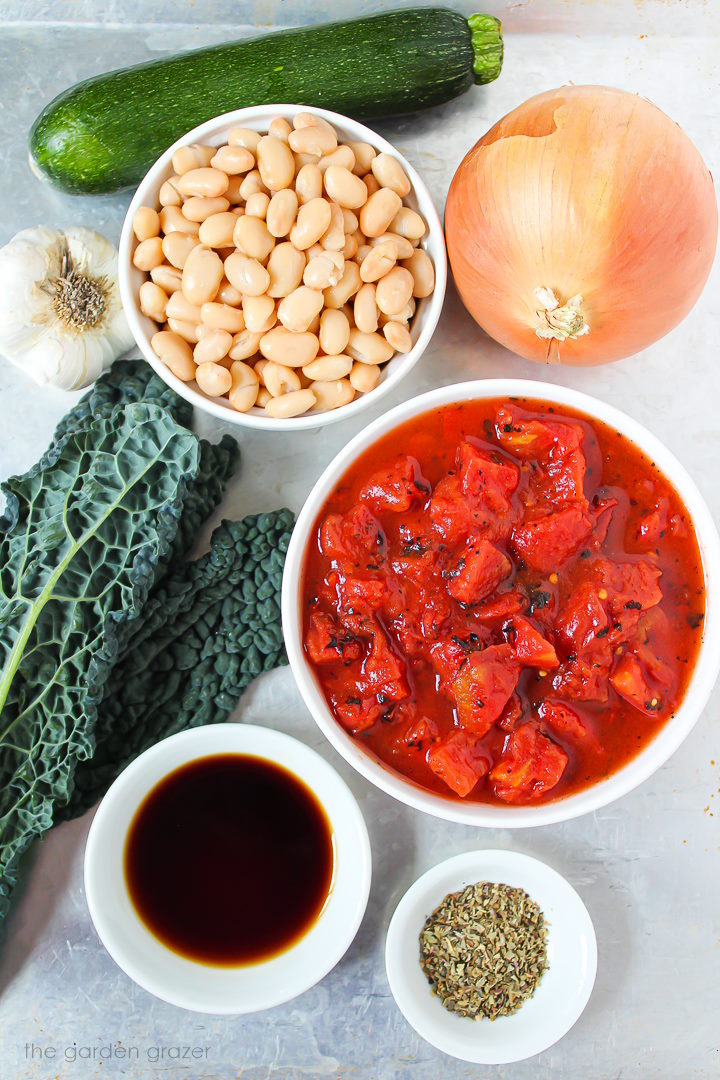 Fresh kale, tomato, bean, onion, garlic, zucchini, and spice ingredients laid out on a metal tray