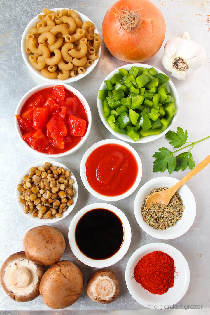 Elbow pasta, mushrooms, lentils, green bell pepper, diced tomatoes, tamari, and seasoning ingredients laid out on a metal tray