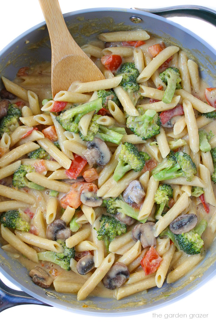 Creamy vegetable pasta with broccoli, mushroom, and tomato cooking in a large skillet with wooden stirring spoon