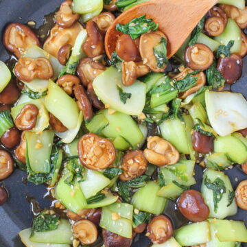 Bok choy and mushrooms cooking in a pan with wooden spoon