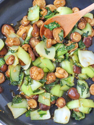 Bok choy and mushrooms cooking in a pan with wooden spoon