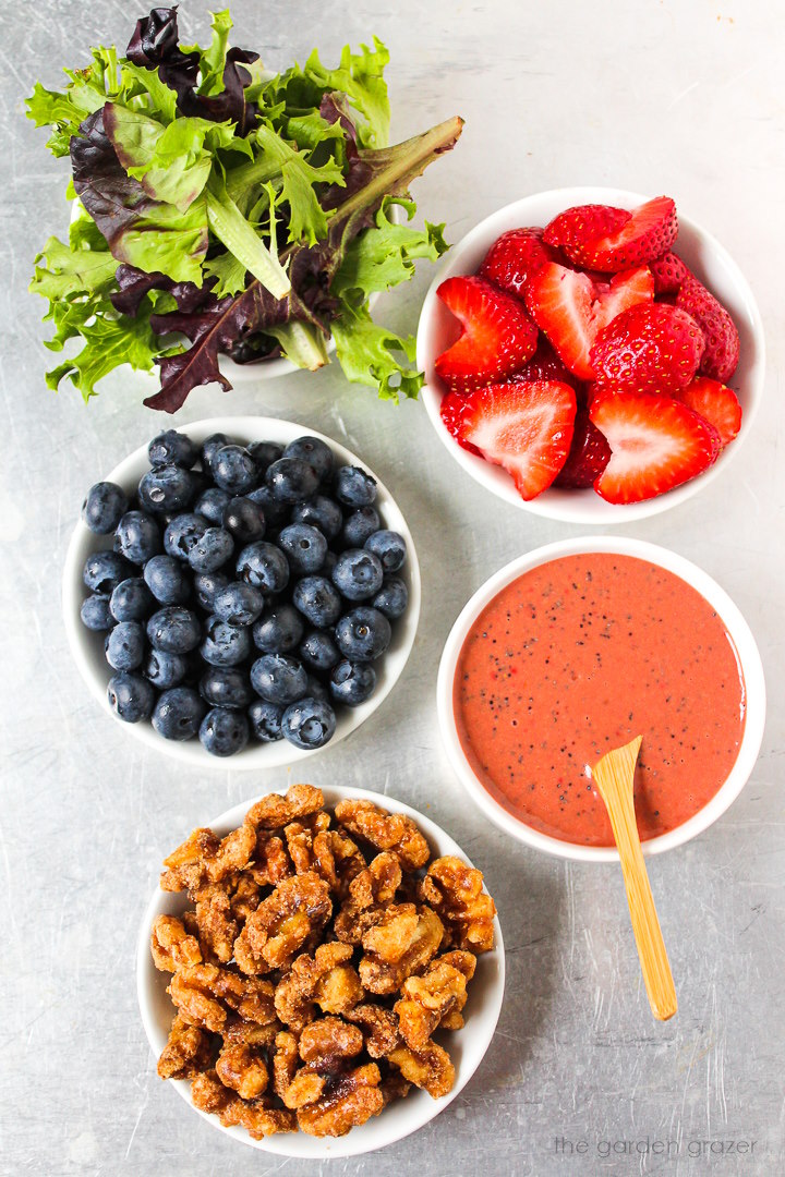 Overhead view of fresh baby greens, fresh berries, candied walnuts, and homemade dressing ingredients laid out on a metal tray