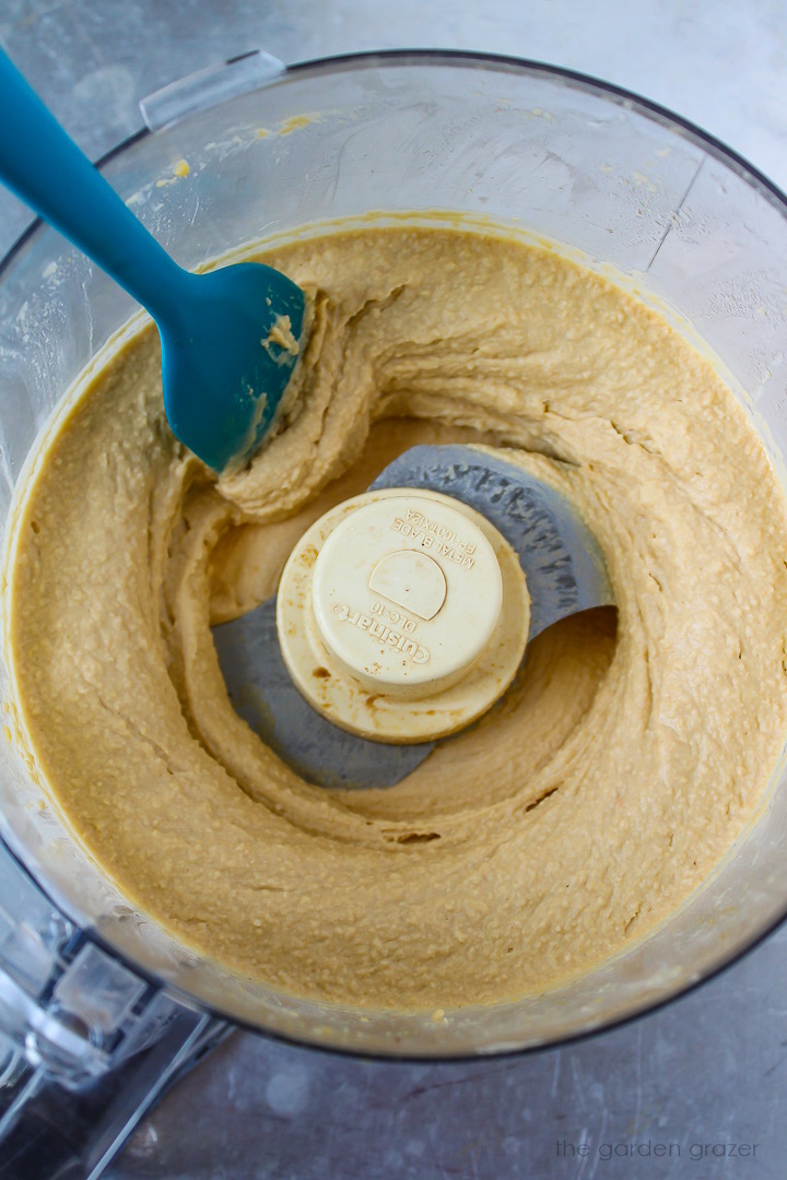 A blue spatula scooping up blended hummus from a food processor