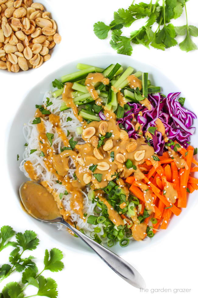 White bowl with rice noodles and fresh vegetables, topped with a peanut sauce