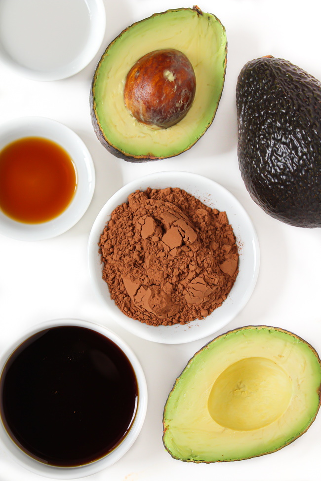 Avocados, cacao powder, maple syrup, and vanilla extract ingredients on a white table