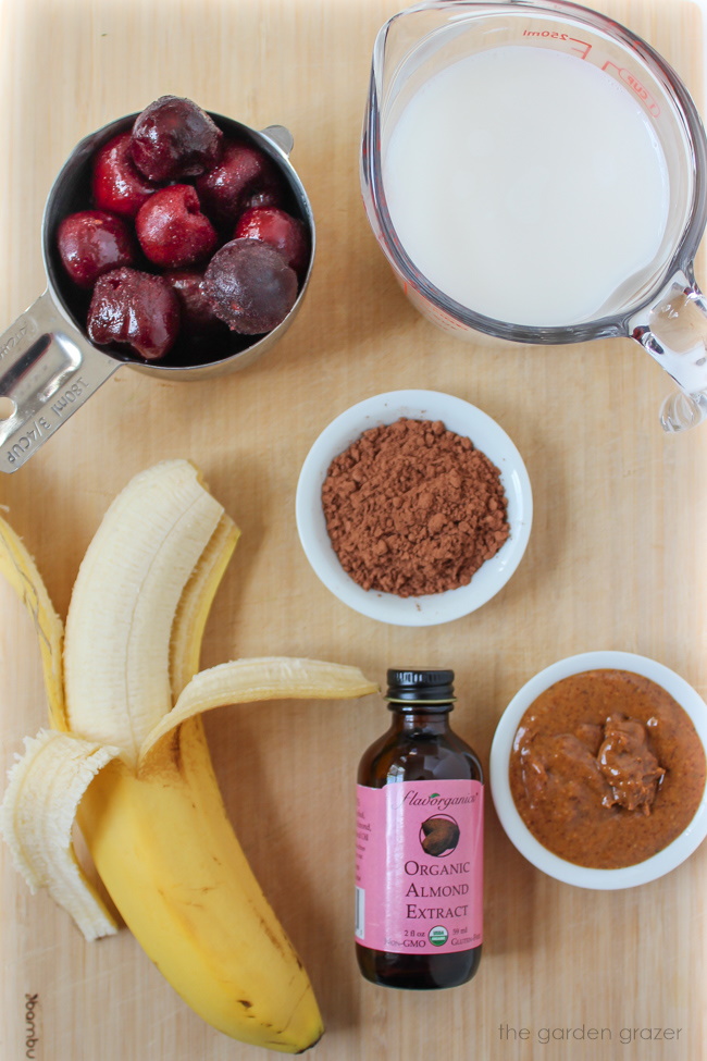 Banana, milk, frozen cherries, and cacao powder ingredients on a bamboo cutting board