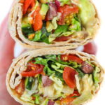 Hands holding a vegan Greek Salad Wrap with hummus cut in half