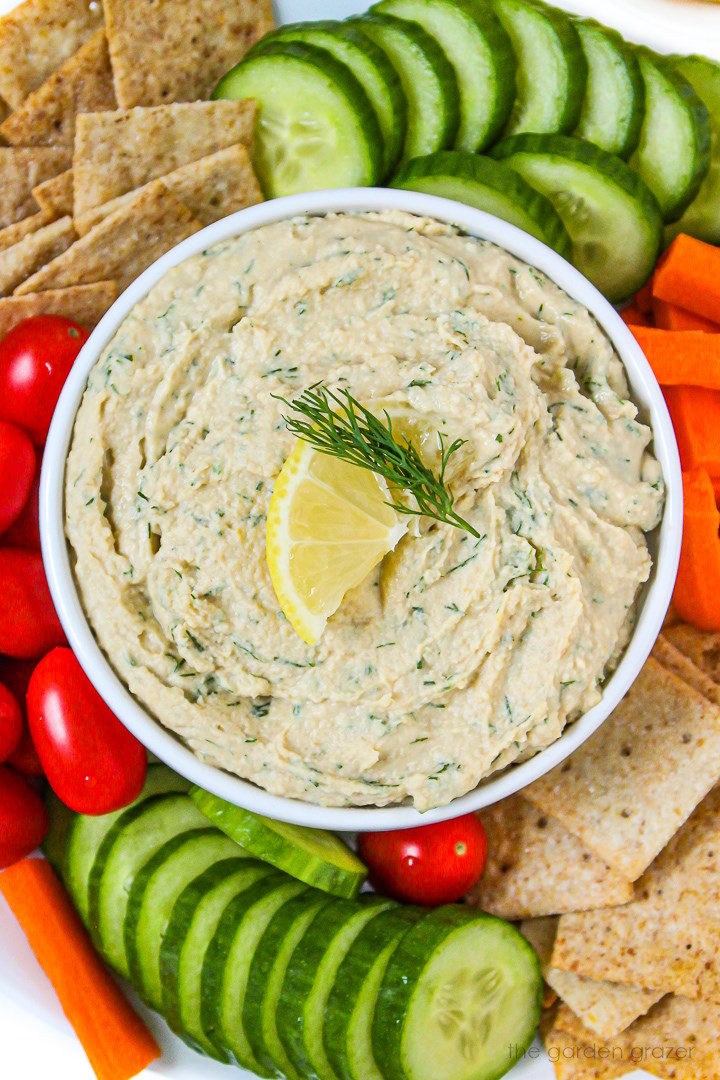 Close up view of lemon dill hummus in a white bowl surrounded by sliced veggies and crackers