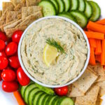 Lemon dill hummus in a white serving bowl with cut veggies and crackers on the side