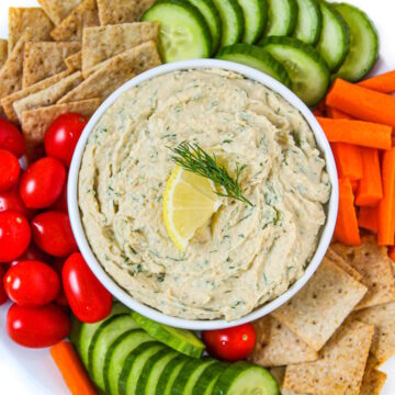 Lemon dill hummus in a white serving bowl with cut veggies and crackers on the side