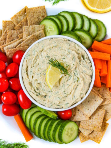 Lemon dill hummus in a white serving bowl with cut veggies and crackers on the side