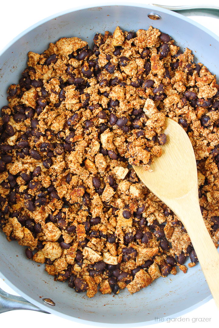 Taco-seasoned tofu and black beans cooking in a skillet with wooden spoon