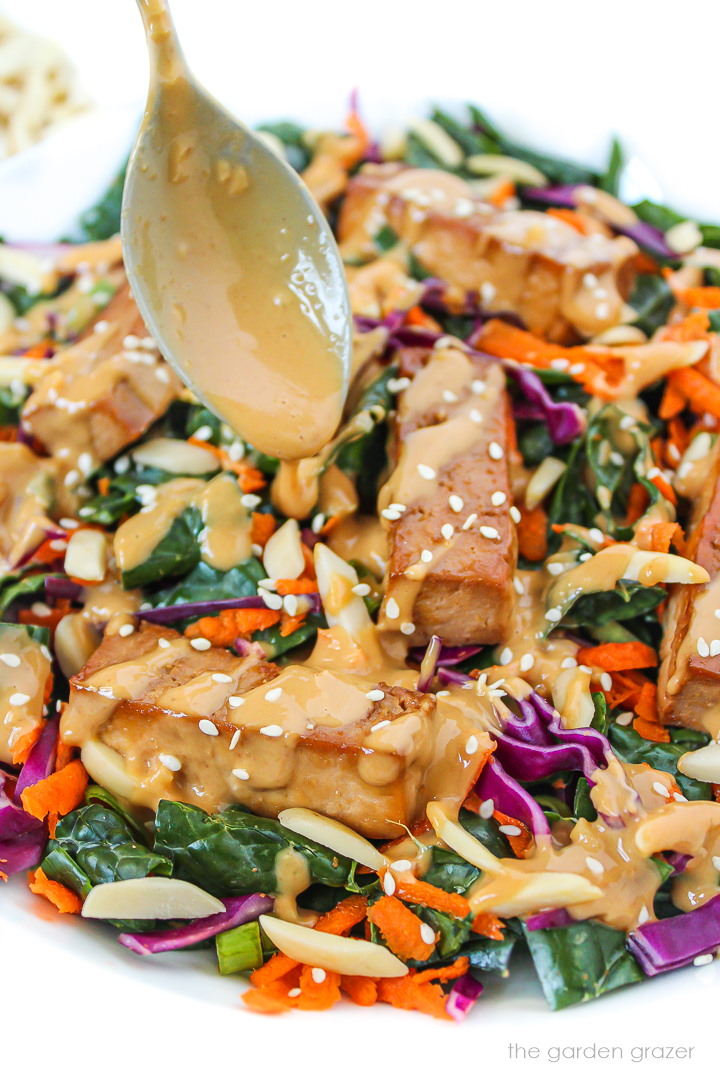 Asian-style kale salad on a white plate topped with baked tofu and creamy peanut dressing
