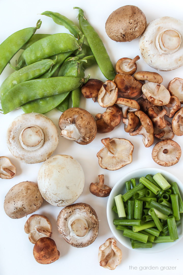 Raw shiitake, white button, and cremini mushrooms with snow peas on a white table