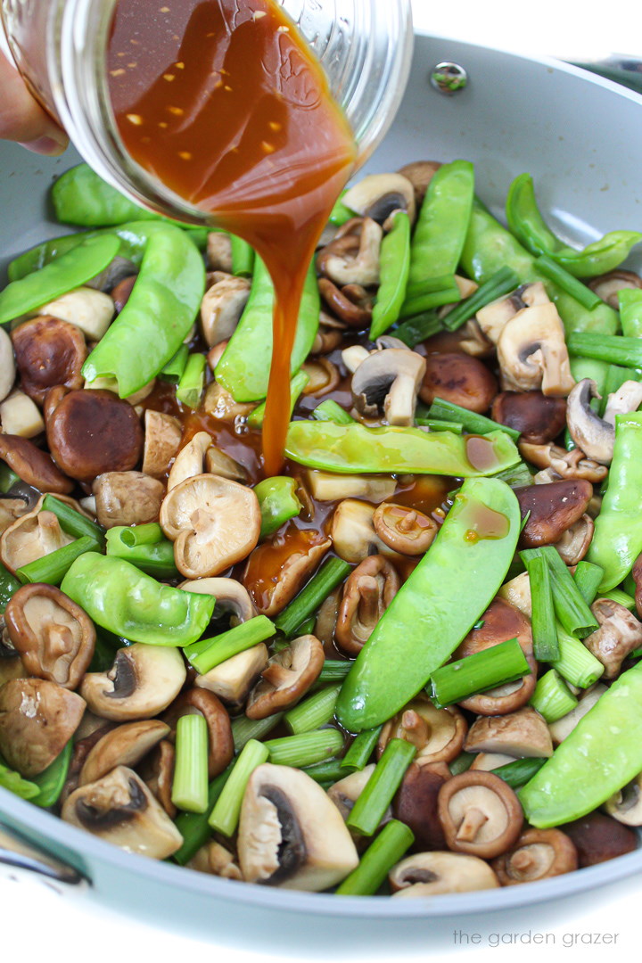 Teriyaki sauce being poured over mushrooms and snow peas in a large skillet