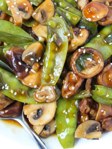 Close-up view of saucy mushrooms and snow peas on a white plate with spoon