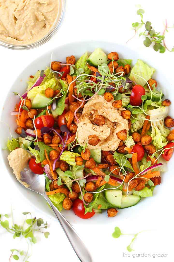 Fresh salad in a white bowl topped with roasted chickpeas, hummus, and balsamic vinegar