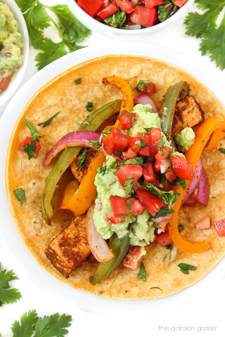 Tofu fajitas on a white plate topped with guacamole and salsa